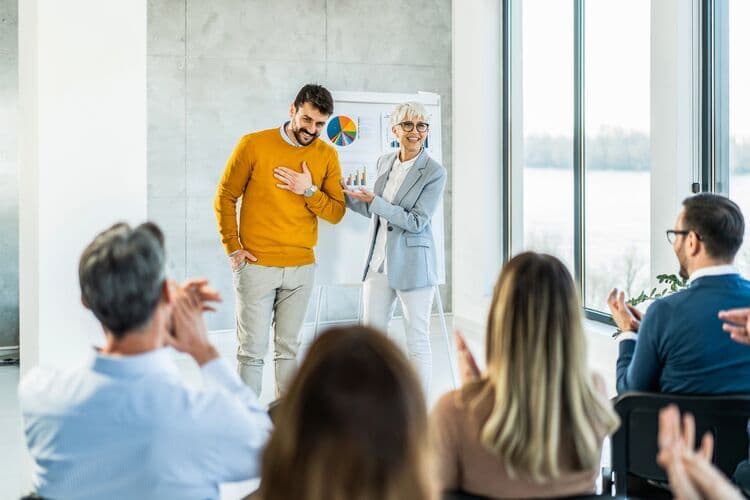 A woman presents her humble colleague to an applauding crowd for appreciation.