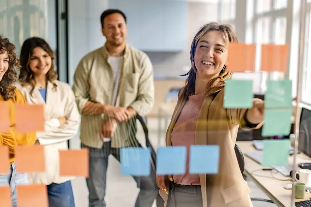 A group of four colleagues is engaged in a brainstorming session in an office setting. A woman in the foreground is smiling while pointing to a sticky note on a glass board, showcasing her enthusiasm. Behind her, three colleagues, one man and two women, are watching with relaxed and supportive expressions, indicating a collaborative environment. The workspace has modern furniture and is well-lit, creating an inviting atmosphere for teamwork.